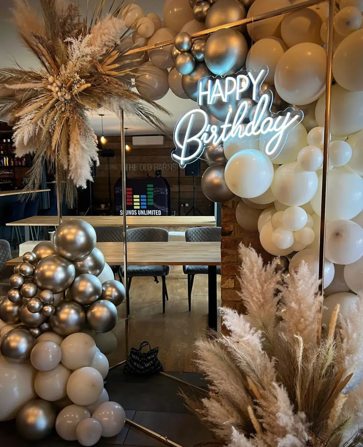 A Happy Birthday neon sign set into an arch of silver and cream balloons with dried pampas grass