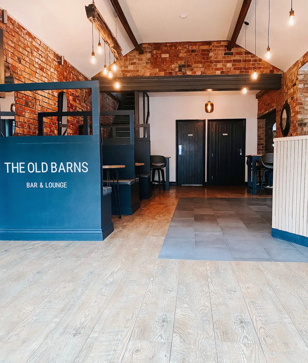 The entrance, looking through to the bar: the Old Barns sign on a slate-blue panel, exposed brick, a timber A-frame and Edison pendants
