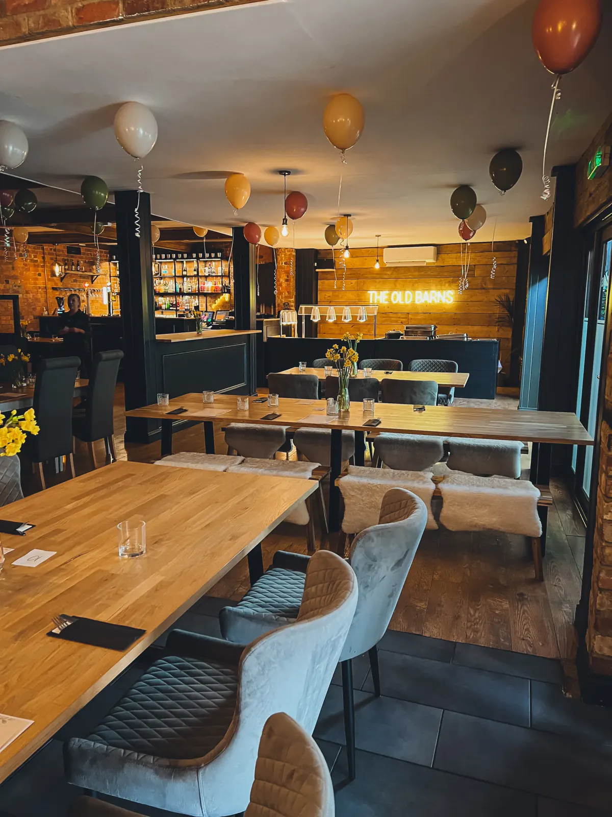 Long oak tables laid out for a birthday buffet, with grey velvet chairs, balloons against the ceiling and the neon sign glowing at the far end of the room