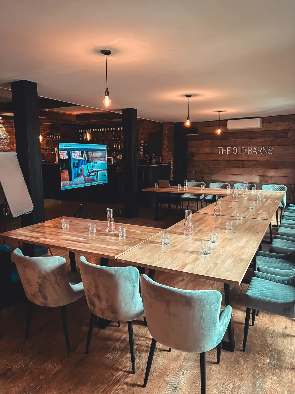 Long oak tables set for a meeting, with teal velvet chairs, water glasses, a screen and a flip chart