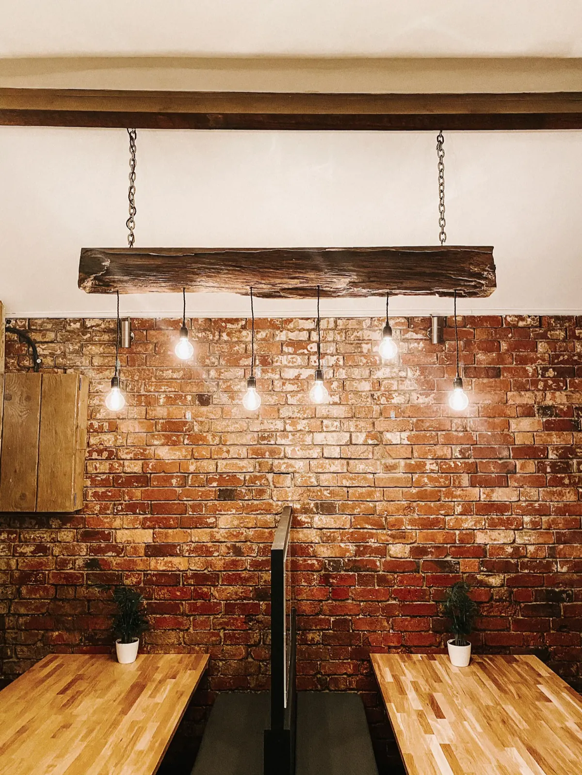 A reclaimed timber beam hung with Edison bulbs above two oak tables against exposed red brick