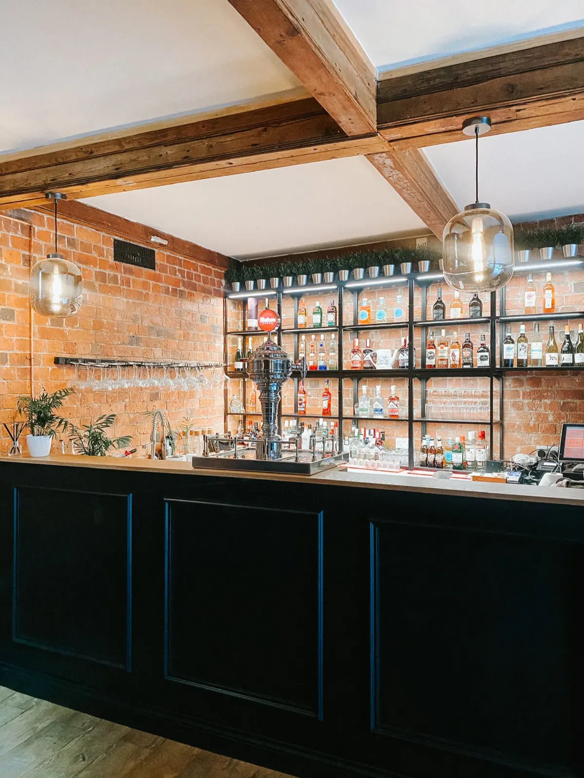 The bar: a dark navy panelled counter under exposed brick and timber beams, with glass pendant lights and back-lit shelves of bottles