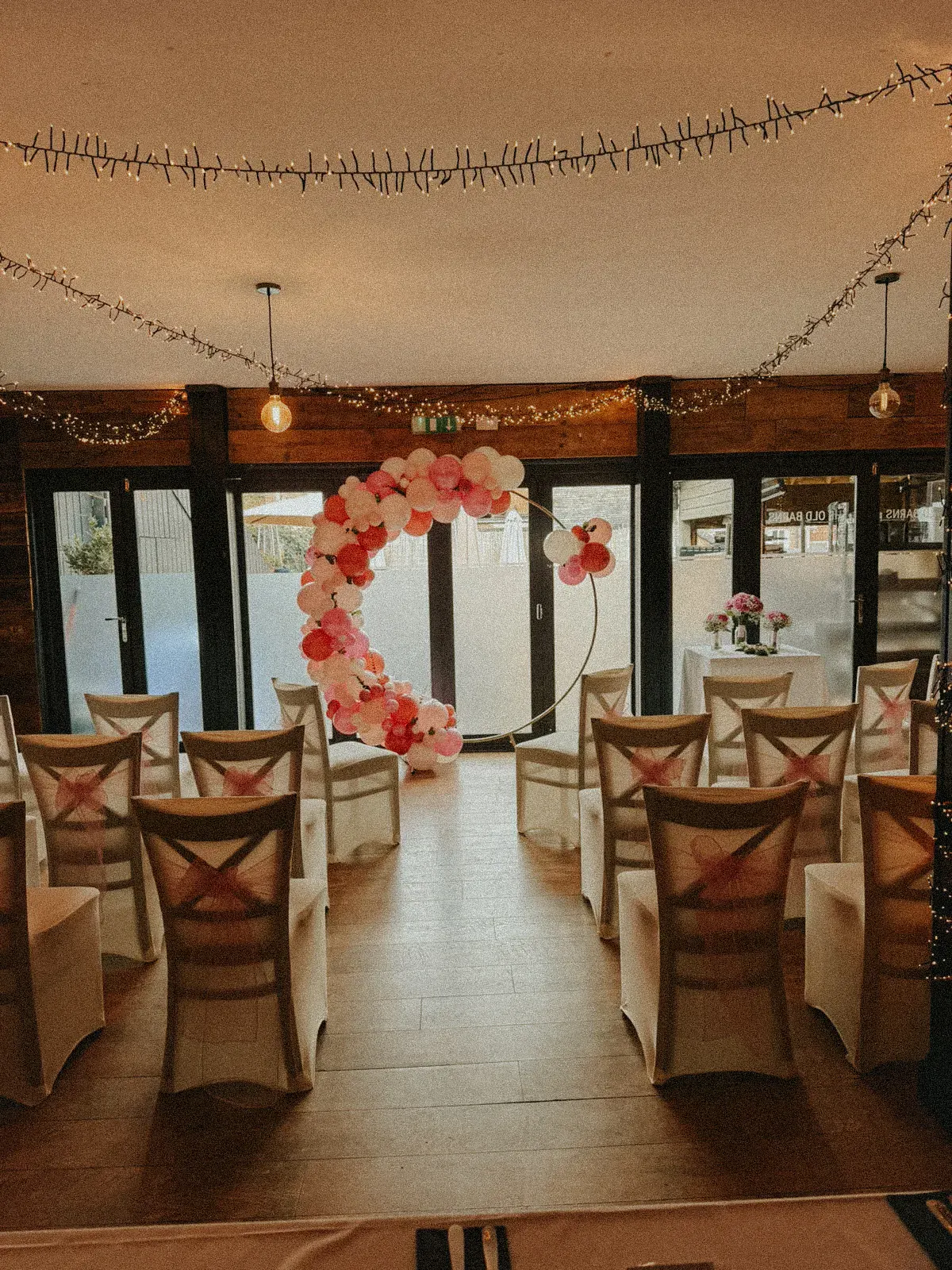 Rows of covered chairs with crossed sashes facing a pink balloon arch, with fairy lights strung across the ceiling and glass doors behind