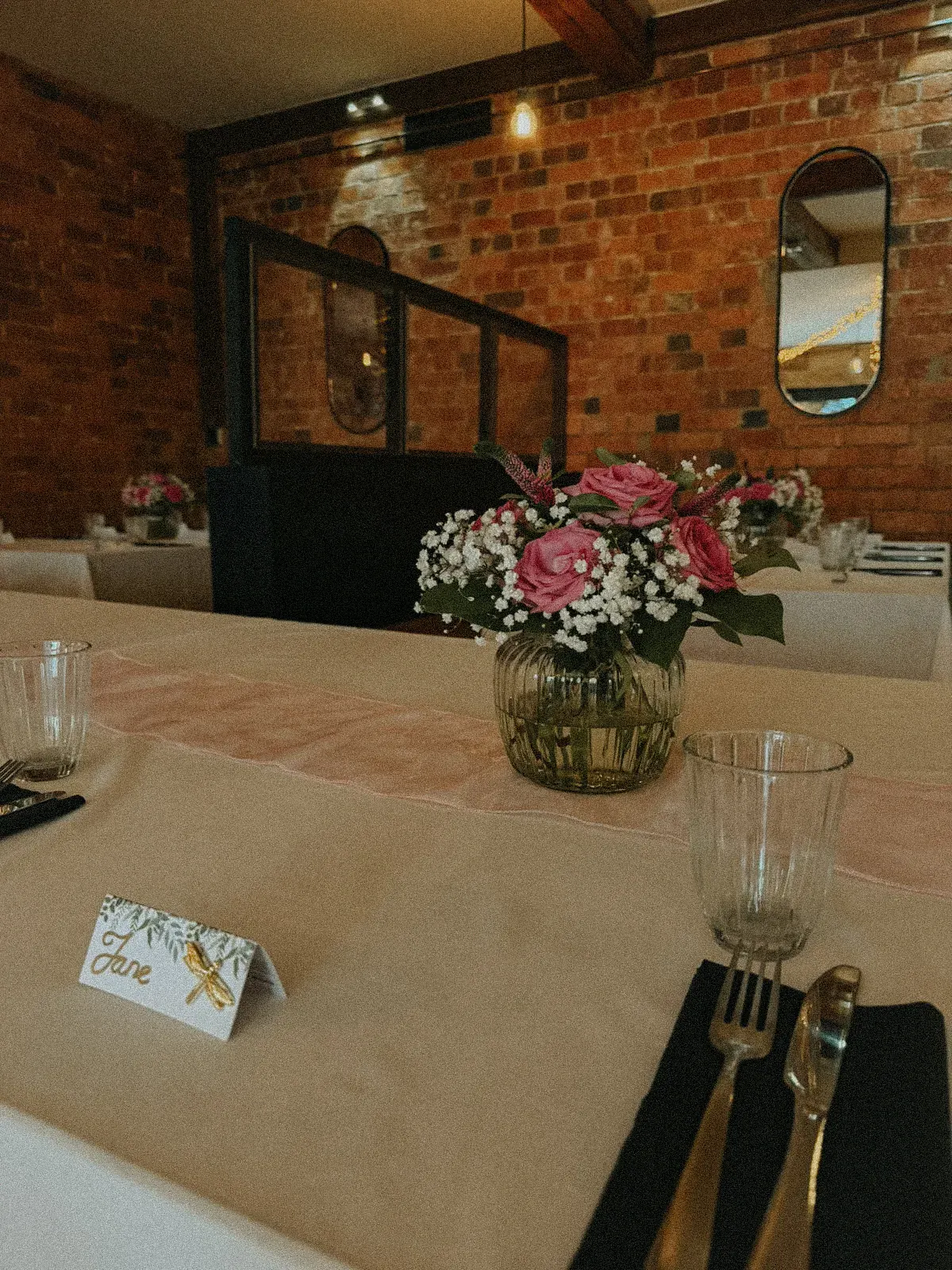 A wedding table laid with white linen, a blush runner, pink roses in a glass vase and a handwritten place card, beneath exposed brick and an arched mirror
