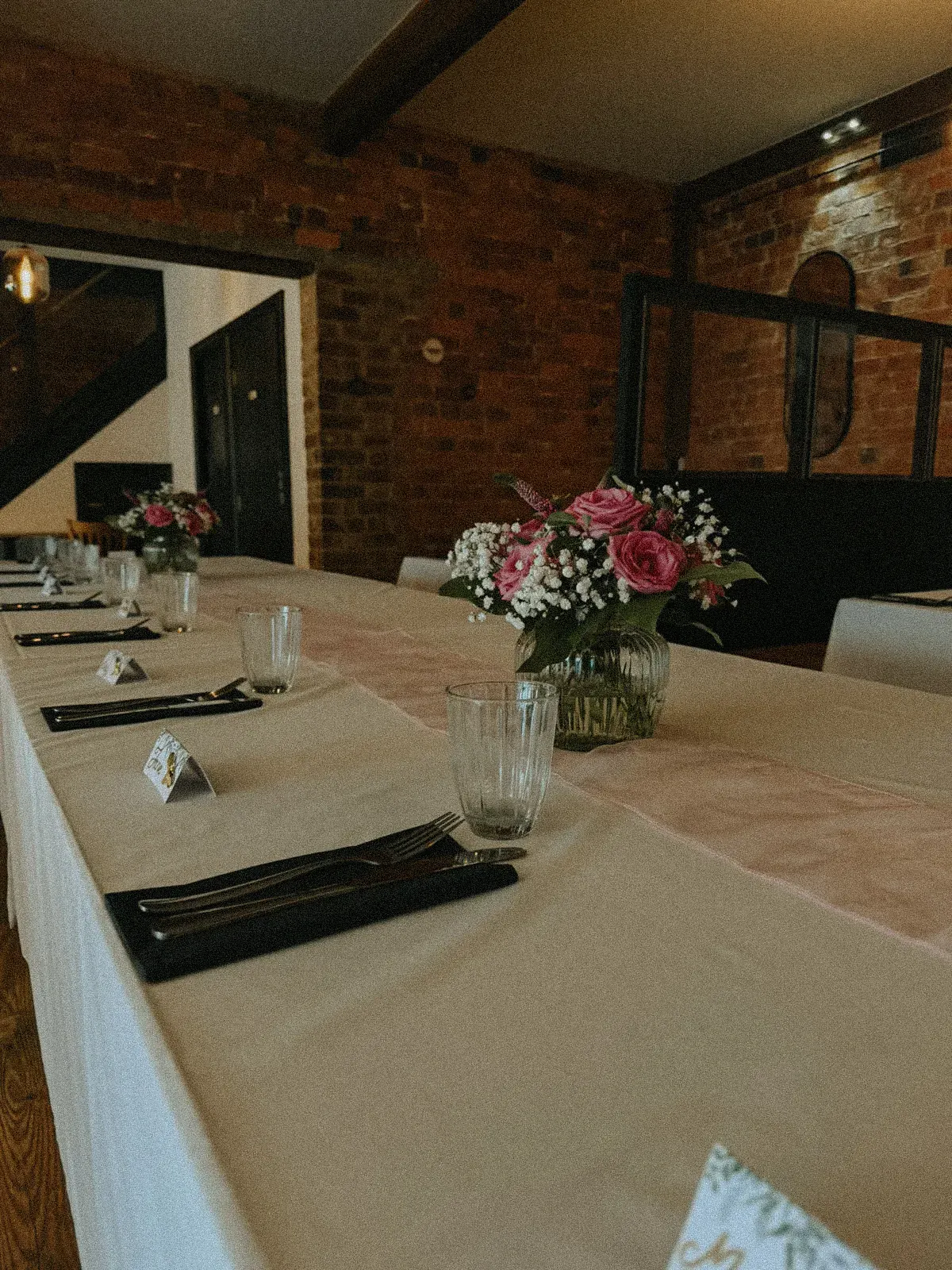 A long wedding table set with white linen, folded black napkins and pink roses, running the length of a brick-walled room